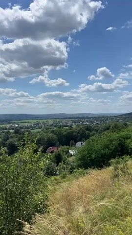 A time-lapse video shows clouds moving across a blue sky above a valley landscape. The valley contains trees, houses, and fields. The foreground is dominated by tall, dry grass and green bushes. The sun is shining brightly, casting shadows.