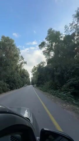 A first-person perspective video shows a car driving on a paved road. The road is lined with lush green trees on both sides, creating a tunnel-like effect. The sky above is a bright blue with scattered white clouds. The car's dashboard and hood are visible in the foreground, along with a rearview mirror. The yellow line in the center of the road indicates it is a two-lane road. There are occasional signs and guardrails visible along the side of the road. The video is taken during daylight hours.