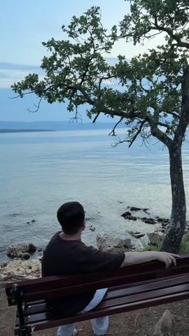 A person is sitting on a bench facing the sea. They are wearing a dark brown t-shirt and white pants. The bench is made of wood and metal. The person is looking out at the water, which is calm and reflects the light blue sky. There are rocks and pebbles along the shoreline, and a tree with green leaves stands to the right of the bench. The scene is set in a peaceful, natural environment, likely near a coast. The time of day appears to be late afternoon or early evening, given the soft lighting and pale sky. The overall mood is one of tranquility and contemplation. The location is identified as Sveti Vid-Miholjice, Croatia.