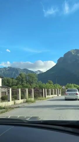 A white car drives down a paved road on a sunny day, with a mountain range in the background. A fence lines the side of the road. The camera is inside another vehicle, presumably the one recording. A red utility vehicle with a driver approaches the camera's vehicle on the road.
