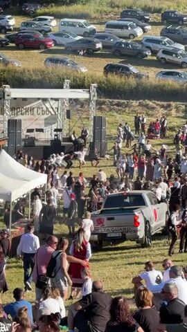 A large outdoor gathering is taking place on a grassy hill with a stage set up on the left side. Cars are parked in rows up the hill behind the crowd. A band is performing on stage, and many people are dancing and mingling in front of it. A white tent is set up to the left of the stage. A silver pickup truck with "DELTA GUARD" written on the side is parked in the middle of the gathering area. The crowd consists of people of various ages, many dressed casually for a summer event. Some individuals appear to be in traditional clothing. The overall atmosphere seems festive.