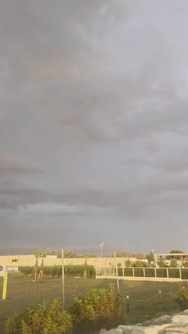 The video displays an overcast sky with dark, ominous clouds. Below the clouds, there is a flat landscape with a grassy field and a few palm trees. There are also some structures that appear to be part of a park or recreational area, including what looks like a volleyball net. The overall atmosphere suggests a gloomy or possibly stormy day.