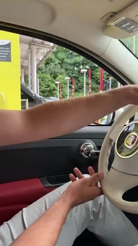 The video begins inside a Fiat 500 car. The driver's hands are visible on the steering wheel and near the gear shift. A passenger's legs are visible in the foreground. The camera pans to show the dashboard, including a mounted smartphone and the car's radio. The scene then shifts to a view from the car's windshield, showing the road ahead. Other cars are visible on the road, and the environment is lined with trees. The sky is cloudy. The car appears to be in motion on a road that may lead to or from a highway.