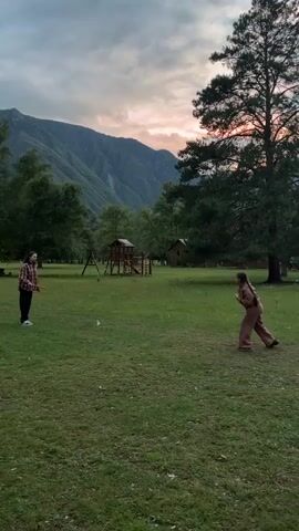 Two individuals are playing a game with racquets and rings on a grassy field. The individual on the left is wearing a plaid shirt and dark pants, holding a blue racquet and a white ring. The individual on the right is wearing a tan tracksuit and is holding a tan racquet and a tan ring. Behind them, a playground structure is visible, along with trees and mountains in the background. The sky is overcast with hints of pink and orange, suggesting dusk. The person on the left attempts to hit a ring with their racquet, while the person on the right waits.