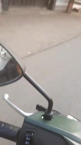 A first-person perspective video shows a person riding a motorcycle. The video begins with a view of a house with a gray roof and white walls. A sign is visible in front of the house that reads "MASJID DESA ME..." and "...KEG. SELO". The rider then moves onto a paved road. Another house with a gray roof and white walls is seen with some potted plants in front. The road continues with some dry, dusty ground on the side. The rider passes by some banana trees with flags attached to poles. The road becomes paved again, and the rider continues forward. A motorcycle with a sidecar carrying a large load of white bags is seen moving in the same direction. Several buildings are visible on the side of the road, and there are other motorcycles parked. The video ends with the motorcycle rider continuing down the road.
