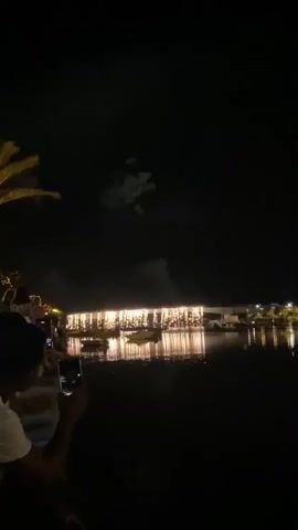 The video captures a nighttime scene with a dark sky, likely in a waterfront setting. In the foreground, a person wearing a white shirt is holding up a mobile phone, seemingly recording something. To the left, a palm tree frond is visible against the dark sky. Across a body of water, a building or structure is illuminated with numerous warm lights, and its reflection shimmers on the dark water. Several boats are docked near the structure. In the sky above the building, there are faint traces of what appear to be fireworks or some sort of aerial display. The overall atmosphere suggests a celebratory event.