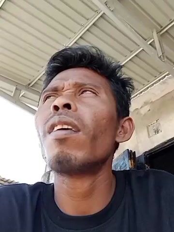 A close-up shot shows a man with dark hair and a goatee looking upwards. He is wearing a black t-shirt. The background appears to be outdoors with a corrugated metal roof above and a textured wall to the right. The man’s facial expression suggests he is thinking or perhaps concerned. The lighting is natural, casting shadows on his face.