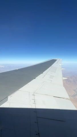 The video shows a view from an airplane window. The primary focus is the airplane wing, which extends diagonally across the frame. The wing is white with some gray sections and appears to be the front portion of the aircraft. The shadow of the wing is cast onto the lower part of the frame. Beyond the wing, a vast landscape unfolds under a clear blue sky. The landscape consists of dry, mountainous terrain with visible valleys and some patches of snow or ice at higher elevations. The horizon is visible in the distance, where the blue of the sky meets the hazy atmosphere above the mountains. The video appears to be taken during the daytime, with the sun casting a distinct shadow from the wing. There are no people visible in the video.
