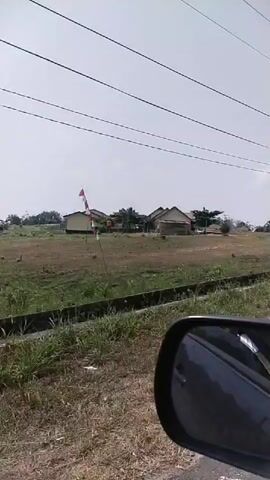 The video appears to be taken from inside a vehicle, looking out the side window and through the side-view mirror. The scene is a rural or semi-rural landscape with a wide, open field. Several small, simple buildings, likely houses, are scattered across the field in the distance. There are also power lines crisscrossing the sky. In the foreground, there is a ditch or drainage channel running parallel to the road, with some grass and vegetation. On the right side of the frame, there are red and white striped poles, possibly marking a railway crossing or some other boundary. Further in the distance, a small building with an orange roof and white walls can be seen, along with what looks like a small gas station. The sky is overcast. The camera is panning or the vehicle is moving slowly, causing the scenery to shift. No people are clearly visible.