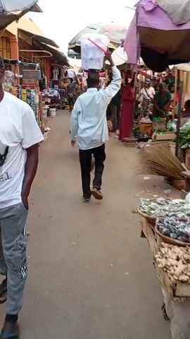 The video shows a bustling outdoor market scene, viewed from a first-person perspective as the camera moves forward along a narrow aisle. The primary focus is on a man in a light-colored long-sleeved shirt and dark trousers, walking away from the camera. He skillfully balances a white plastic bucket on his head, occasionally steadying it with his right hand.

On the left side of the aisle, there are numerous market stalls. These stalls display a variety of goods, including shelves stacked high with colorful packaged items like snacks and possibly drinks. Further down, large woven sacks, possibly containing dried beans or similar agricultural produce, are placed directly on the ground. Some of these sacks have text visible, reading "ASS MUDA." Baskets filled with red onions are also seen on this side.

On the right side of the aisle, more market stalls are visible, covered by large, colorful umbrellas providing shade. Several other people are walking and standing in the market, including vendors and shoppers. One woman in a striking red dress is particularly noticeable among the crowd. The ground appears to be a paved surface, likely concrete or asphalt. The lighting suggests it is daytime, possibly morning or afternoon, with ample natural light filtering through the covered stalls. The overall atmosphere is that of a busy and active local market.