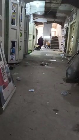 The video depicts a poorly maintained interior space, likely a corridor or covered alleyway, with a concrete floor littered with debris including plastic bottles and paper. The lighting is dim, and the area appears somewhat grimy.  On the left, there are structures resembling large lockers or storage units, painted in a light color with contrasting trim.  To the right are closed doors.  Two figures are visible.  One, further in the background, is wearing a dark robe.  The other, closer to the camera, is dressed in a faded green top and khaki pants. This second person is carrying a bundle of goods on their head and dragging a large, possibly filled, plastic bag along the floor.  They are walking barefoot. A set of stairs is visible in the background.