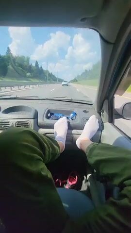 The video shows the interior of a car from the passenger's perspective. A person's bare feet in white socks are resting on the dashboard. In the background, through the windshield, a highway is visible with cars driving in both directions. The sky is blue with white clouds, and trees line the sides of the road. The car appears to be in motion.