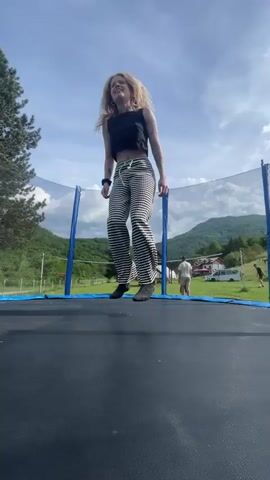 A woman with blond hair wearing a black tank top and black-and-white striped pants is jumping on a trampoline.  The trampoline has a blue frame and a safety net. The woman falls onto her seat and then stands back up. The background features green hills, trees, and a partly cloudy sky. In the distance, there are a few buildings and people can be seen moving around.