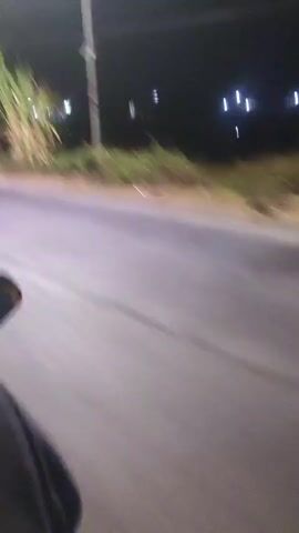 The video shows a blurry, night-time view from a moving vehicle, possibly a motorcycle, looking towards the right side of the road. In the foreground, a portion of the vehicle's body and a rider's leg are visible. The road appears to be asphalt. On the right side of the road, there is a grassy area with some tall plants and foliage. In the background, streetlights are visible, casting a glow in the darkness. The overall impression is of a journey through a rural or semi-rural area at night.