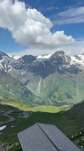 A stunning panoramic view of snow-capped mountains under a bright blue sky with fluffy white clouds. The foreground features a dark-roofed building and a flagpole with a red and white flag, likely the Austrian flag. Lush green valleys and slopes with winding roads are visible in the midground. The scene appears to be captured from a high vantage point, offering a breathtaking natural landscape.
