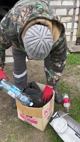 The video shows a person wearing a camouflage jacket, gray pants, a gray striped beanie, and red gloves leaning over a cardboard box on a grassy surface. The box appears to contain various items including a black bag, orange string, gloves, and other miscellaneous objects. Two water bottles lie on the ground beside the box, along with ear protection, a white light fixture, and reflective foil. The background includes a building with white siding and a glimpse of a vehicle. The person is digging through the box.