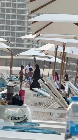 The video pans across a busy beach with numerous lounge chairs and umbrellas. In the background, a tall, modern building stands. Several people are visible, including a woman in a black abaya walking between lounge chairs, two children playing near the water, and other individuals relaxing on the beach. In the foreground, a small white table holds various items, including a bottle of soda, a red cup, a bag of chips, and a towel. The overall scene depicts a lively day at the beach.
