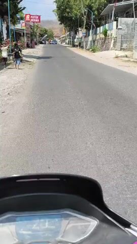 The video shows a POV shot from a motorcycle. The motorcycle is traveling down a paved road in a town. On the left side of the road are buildings with shops and awnings. People are walking on the sidewalk. On the right side of the road are buildings and a wall. There are cars and a bus on the road. The sky is clear and it is daytime. The dashboard of the motorcycle shows the speed as 20 km/h.