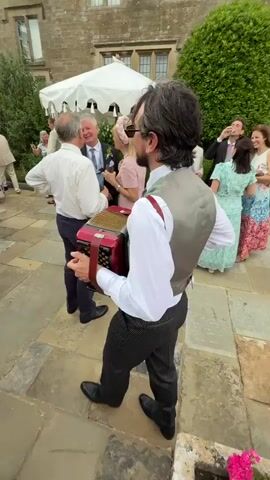 This video captures a lively outdoor gathering at a large stone building, possibly a manor or castle, located in the Cotswold District, United Kingdom. The scene suggests a daytime event, likely a wedding or celebratory party, with guests dressed in formal attire. The weather appears pleasant, with white canopies providing shade on the stone-paved courtyard.

The focal point of the video is a man playing an accordion, dressed in a white shirt, grey vest, and pinstripe trousers. He is positioned in the foreground, facing away from the camera, and appears to be entertaining a small group of guests. To his right, a man in a white shirt and dark suit is talking with a woman in a pink dress and a decorative headband. Another man, also in a suit, stands nearby, holding a dark jacket. Several other guests are visible in the background, mingling and enjoying the occasion.

The architecture of the building, with its stone facade and multiple windows, suggests a historic and grand setting. The presence of large white parasols adds a festive touch to the outdoor space. The overall mood seems joyful and social, with people engaged in conversation and entertainment. No visible text is present in the video.