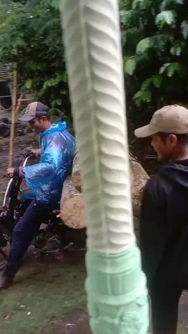 A man wearing a blue and white plastic raincoat, a baseball cap, jeans, and brown shoes is starting up a motorcycle loaded with logs in a yard. The motorcycle is black and has about seven round logs strapped to its back. Another man wearing a grey baseball cap and a blue and grey raincoat is crouched down in the foreground, drinking from a cup. Behind the men is a tan house with white trim. Another motorcycle is parked near the house. The yard is green but wet with mud. A white bucket with "NIPPON PAINT" printed on it sits near the crouched man. Trees and other houses are visible in the background. The camera is filming from under a covered porch.