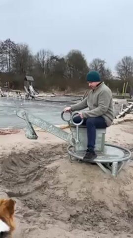 Here is a description of the video:

The video shows a man sitting on a metal playground excavator toy in a sandy area. He is wearing a light green jacket, dark pants, and a dark green beanie. He smiles as he turns the excavator. A dog, with brown, black, and white fur, walks in front of the excavator. The background shows a playground area with trees and other playground equipment, some of which is covered in ice. The sky is overcast. The dog barks at the man.
