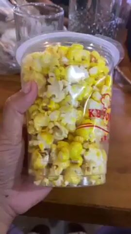 A hand holds up a clear plastic cup filled with popcorn, some of which is colored yellow. The cup has a clear plastic lid and a red, white, and yellow logo that reads "Ke' Popcorn Since 2020". There are three red stars under the logo. The popcorn appears to be a mix of traditional white popcorn and a yellow-colored variety. In the background, there are clear glasses on a wooden surface, and other blurry objects suggest a table setting in a cafe or restaurant. The video is taken at a close range.