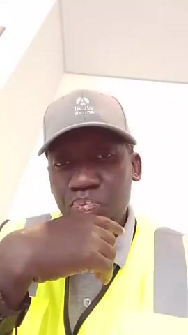 A man wearing a gray baseball cap and a yellow safety vest is in a room with white walls. He makes a gesture with his hand, moving his finger across his cheek and then up to his nose, before moving his hand to his chin and mouth. He is looking directly at the camera.