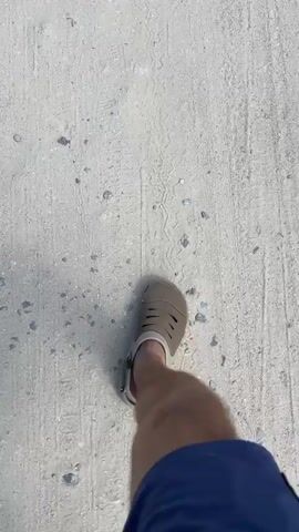 A first-person perspective video shows a person walking on a light-colored, rough surface that appears to be dirt or gravel. The person is wearing blue shorts and brown sandals. The camera angle is downward, focusing on the person's legs and feet as they walk. The surface has tire tracks and small stones scattered across it. The lighting suggests it is daytime.