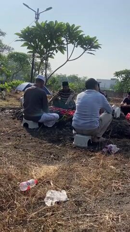 Several men are gathered around a gravesite. They are sitting on small white stools. The grave is decorated with red flowers. There is trash on the ground, including a plastic water bottle and plastic wrappers. There are trees and a street light visible in the background. The sky is blue and the sun is shining.