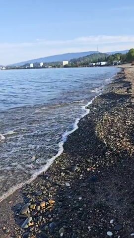 The video features a static, eye-level shot of a calm pebble beach on a clear, sunny day. Gentle, small waves lap the shoreline, which is composed of dark, varied-sized pebbles and some sand. In the background, across the expanse of blue water, a distant coastline is visible, featuring green hills or mountains, several buildings, and a thin tower or antenna on a hilltop. The sky above is a bright, cloudless blue. No people or animals are visible in the frame.