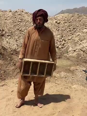 In an arid, sun-drenched outdoor setting in Deh-E Bagh, Afghanistan, a man stands facing the camera. He is dressed in a traditional loose-fitting brown tunic and matching pants, with a dark red turban wrapped around his head. He has a beard and is barefoot.

The man holds a wooden mold at waist level, which contains four rectangular compartments. These compartments appear to hold semi-formed bricks or mud blocks, indicating a brick-making or construction-related activity.

Behind him, large mounds of light brown dirt and rubble dominate the landscape, suggesting an excavation site, a quarry, or an area where building materials are being processed. To the man's right, a small, shallow pool of muddy water is visible on the ground. The sky above is bright and clear, indicative of a sunny day, likely midday given the strong light. The man looks directly at the camera, seemingly speaking, though no text is visible.
