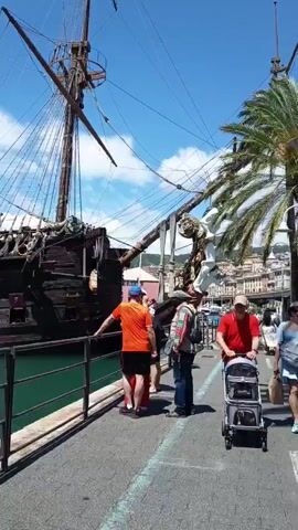 This video captures a scene at a port in Genoa, Italy, on a bright, sunny day with a clear blue sky and some scattered white clouds. The main subject is a large, elaborate wooden sailing ship, appearing to be a replica of a galleon or pirate ship. It features multiple masts, extensive rigging, and intricate gold-colored carvings and decorations, particularly visible on its stern. A red flag flies from one of its masts.

Several people are visible on a concrete pier or dock next to the ship. In the initial frames, a man in an orange shirt leans on a black metal railing, looking towards the vessel. Other individuals are also present, observing the ship or walking along the dock. Later, a young boy in a patterned shirt stands by the railing, also looking towards the ship.

The ship is docked in a body of green-tinged water, typical of a harbor. Smaller boats, including an inflatable dinghy with an outboard motor (where the brand name "HONDA" is visible), are moored nearby. In the background, across the water, various buildings with different colored facades can be seen, contributing to the urban port atmosphere. The overall impression is one of a tourist attraction or a historical exhibit at a vibrant waterfront location.