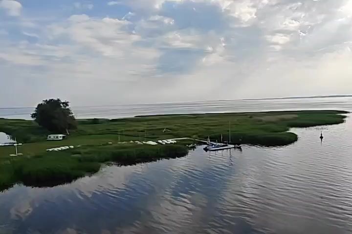 A drone shot flies over a body of water, showing a narrow strip of land with green grass and reeds. On the land, there are several small white sailboats lined up, a pier with a few boats docked, and a white recreational vehicle. The water is calm and reflects the sky, which is partly cloudy. In the distance, a small motorboat is moving across the water, leaving a wake behind it. There are a few buoys in the water.