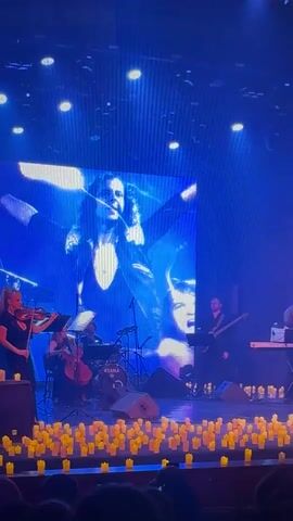 A musical ensemble performs on a stage lit with blue and white lights. The band includes two violinists, a cellist, a drummer with a "TAMA" drum kit, a bassist/guitarist, and a keyboardist. Behind the musicians, a large LED screen displays abstract blue and white visuals. The front of the stage is adorned with numerous yellow candles, creating an intimate ambiance. The musicians are actively playing their instruments, and an audience is partially visible in the foreground, watching the performance. The scene depicts a live concert or performance indoors.