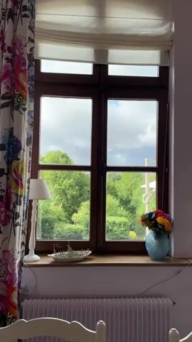 The video shows a close-up, stationary shot of a wooden-framed window with two panes. Outside the window, lush green trees and foliage are visible under a cloudy sky. The windowsill is decorated with a white lamp on the left, a decorative dish with dried sprigs in the center, and a blue vase filled with colorful flowers on the right. To the left of the window, a floral curtain with large pink, blue, and yellow flowers is partially visible. At the bottom of the frame, a portion of a white radiator and the top of a white chair with a carved back are in focus. The overall scene suggests a bright, natural-lit indoor space overlooking a garden.