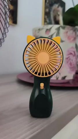 A close-up, static shot shows a dark green handheld fan with yellow cat ears and a yellow fan cover sitting on a light wooden surface. The fan's blades are visible through the cover and are spinning. In the background, a pink decorative plate is visible, and behind that, a large ceramic pot with a floral pattern of pink and white flowers. Further in the background, two framed pictures hang on a wall, with one appearing to be a nature scene and the other a picture of an animal. The lighting suggests an indoor setting, possibly a room with natural light. There are no people or interactions visible.