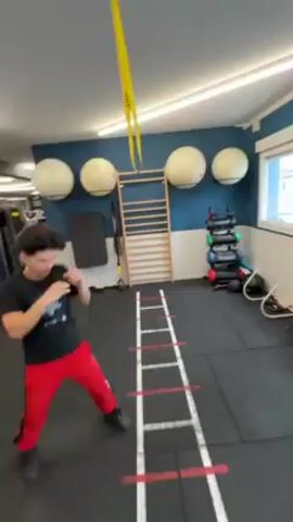 A young man is doing a footwork exercise in a gym. He is wearing a black t-shirt with a graphic on it, red pants with a black stripe, and black shoes. He is standing on a black floor with a red and white agility ladder painted on it. He is moving his feet quickly and sideways between the rungs of the ladder, while holding his hands up in a boxing stance. The gym has blue walls with white trim. There is a window, a ladder, white sphere-shaped decorations, a weight rack, and a TV on the wall. The TV is showing workout content. There is also a yellow TRX suspension trainer hanging from the ceiling. The room is well-lit with fluorescent lights.