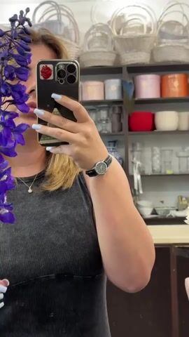 A medium shot shows a person with blonde hair holding a smartphone in their right hand, taking a selfie in a mirror. The person is wearing a grey tank top and a black watch on their left wrist. Their fingernails are painted light blue. A bunch of purple delphinium flowers are held in the person's left hand, partially obscuring their face. In the background, shelves are filled with various items, including white baskets, colorful gift boxes, and glass vases. The person's arm is visible on the right side of the frame. The environment appears to be a shop.