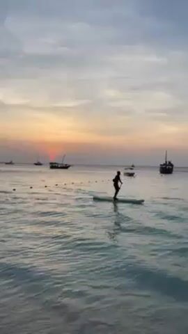 The video shows a person paddleboarding in a calm, turquoise ocean at sunset.  The sky is a mix of light blues, grays, yellows, and oranges. The sun is partially obscured by the horizon, casting a vibrant orange glow on the water. Several boats are visible in the distance, silhouetted against the setting sun. The person on the paddleboard is also silhouetted and appears to be paddling away from the camera. There are floating buoys in the water nearby. The overall scene conveys a sense of peace and tranquility.