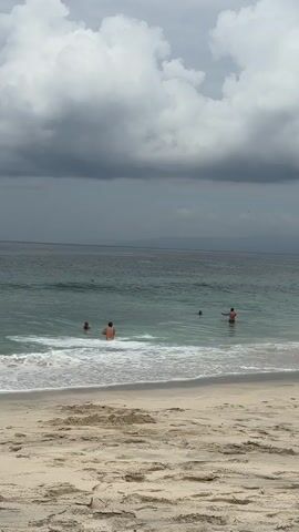 This is a short video clip of a beach scene in Manggis, Indonesia. The foreground shows a sandy beach with subtle ripples in the sand. The middle ground features the ocean with gentle waves breaking on the shore. Several people are visible in the water, some appearing to be swimming or wading. The water is a clear turquoise color near the shore and transitions to a deeper blue further out.

The background depicts a vast expanse of the ocean meeting the horizon, with a hazy line of land visible in the distance. The sky is dramatic, filled with large, dark storm clouds that contrast with patches of brighter sky. This suggests a somewhat overcast or potentially rainy day, perhaps late afternoon or early evening when the light is diffused.

The overall mood of the scene is serene and relaxed, despite the cloudy sky. The presence of people enjoying the water adds to a sense of leisure and vacation. There is no visible text in the video clip. The focus is on the natural beauty of the beach and the activity of people enjoying the ocean.