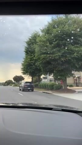 The video shows a view from inside a vehicle driving down a road. A black SUV is in front of the vehicle, traveling in the same direction. The road has multiple lanes and is lined with trees and some buildings. The sky is cloudy, and there appears to be rain in the distance. The windshield of the vehicle has water droplets on it.