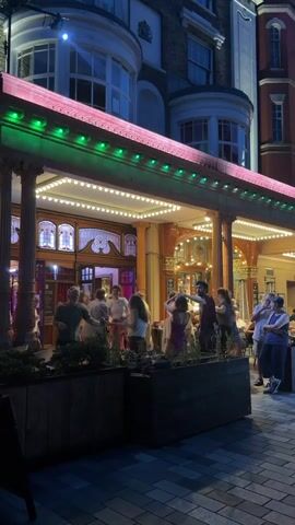The video shows a nighttime exterior scene of a building with a covered outdoor seating area. There are plants in wooden planters in the foreground. A group of people are dancing in the covered area, which is illuminated by strings of lights. Other people are standing and watching. The building facade appears ornate, with columns and decorative windows. The overall atmosphere seems lively and social.