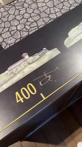 The video displays a tabletop display with a timeline graphic. The graphic depicts a historical building at various stages of its existence, indicated by numerical timestamps. The timestamps visible include 1600, 1820, and 2018. In addition to the graphic, there are three-dimensional white models of the building, placed on different sections of the timeline. The display is on a table with a patterned surface and wooden flooring is visible around the table. One person's feet, wearing leopard-print shoes, are visible at the bottom of the frame. The camera moves across the display, capturing different parts of the timeline and the building models.
