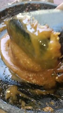 A close-up shot shows a thick, brown sauce being poured from a ladle into a dark, textured bowl or mortar. The sauce has a slightly granular appearance and is glistening. Some of the sauce has spread out in the bowl, creating an oily sheen. A dark, cylindrical object, likely a pestle, is partially submerged in the sauce, suggesting it is being used to grind or mix the ingredients. The background is out of focus, but appears to be a light-colored, rough surface, possibly outdoors. The lighting is natural and highlights the texture of the sauce and the bowl.