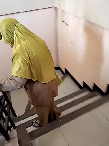A person wearing a yellow hijab and a brown outfit is walking up a set of stairs. The person is holding onto a black railing on the left side of the stairs. The stairs have brown treads and white risers. There are several framed pictures on the wall to the left of the stairs. The wall is a pale pink color. There is a black electrical outlet on the wall to the right of the stairs. The lighting in the scene is dim.