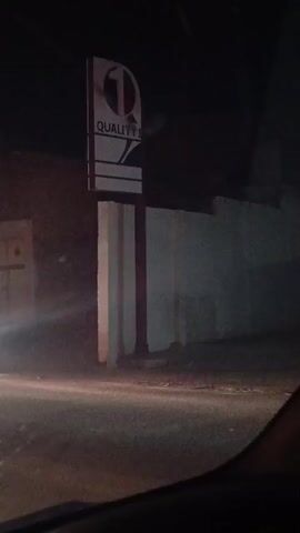 A car is driving past a Quality 1 sign at night. The sign is illuminated and features a "Q1" logo, along with white stripes on a black background. The car's headlights shine on the sign as it passes. Other cars and buildings can be seen in the background, but the focus is on the sign as the car passes. The video appears to have been filmed from inside the car.