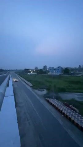 The image presents an elevated view of a road system at dusk or twilight, likely from a bridge or overpass. A white concrete barrier with a railing frames the left side of the composition, extending into the distance. Below and to the right, a multi-lane asphalt road stretches away from the viewer, appearing mostly empty. A single red or orange vehicle with illuminated headlights is visible on this lower road, moving away.

Adjacent to the right side of the lower road, a distinct barrier featuring a yellow and black checkered pattern is present, possibly serving as a safety or demarcation structure. Beyond the roads, the landscape consists of expansive green fields and grassy areas, hinting at a semi-rural or developing urban periphery.

In the far distance, several low-rise to mid-rise buildings are scattered, some displaying illuminated windows or surrounded by streetlights, indicating human activity. The sky is a soft, muted blue-grey, characteristic of the "blue hour," with no direct sunlight visible, suggesting an overcast evening or early morning. The overall ambient light is dim, with artificial lights beginning to stand out.

The scene depicts a quiet and tranquil moment with minimal activity. Based on the provided context, the location is Rupganj, Bangladesh. There is no visible text in the image.