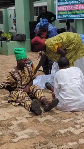 The video shows an outdoor scene with several people gathered. In the foreground, two individuals are sitting on a paved surface, dressed in traditional clothing. One person, wearing a green headscarf and a patterned tunic and pants, is smiling and looking towards the left. Another person, wearing a red head covering and a yellow and green patterned garment, is leaning down and interacting with the first person. Behind them, another person in a white robe is kneeling. In the background, there is a building with green walls and a stone facade. A banner is displayed on the building, featuring text related to a flag-off ceremony. Other individuals are visible in the background, some standing and some seated. The environment appears to be a courtyard or open area outside a public building, possibly for an event or ceremony.
