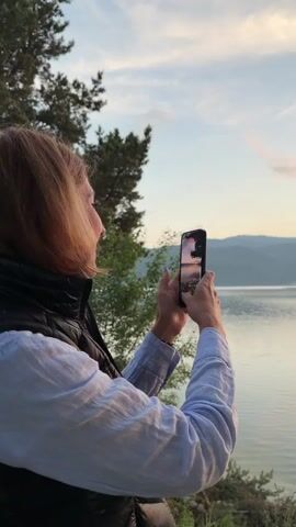 A person stands on the shore of a lake, recording the sunset with a phone. The person then lowers the phone, and the shot focuses on the sunset over the lake. The sun is setting behind mountains, creating a vibrant orange reflection on the water. Pine tree branches hang into the frame from the top.