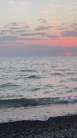 The video shows a peaceful sunset scene at a beach. The sky is filled with pastel clouds in shades of pink and purple, with the sun partially visible on the horizon, casting a warm glow. Gentle waves lap against the pebbled shore. Two people are visible in the water, appearing to be swimming or wading as the sun sets. The overall atmosphere is serene and tranquil.